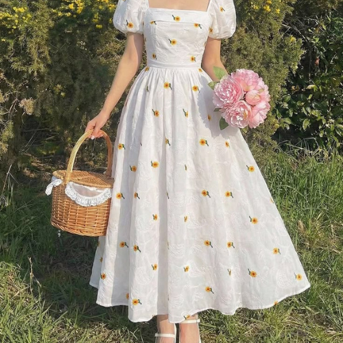 A woman wearing a white midi dress with yellow and white daisy floral prints stands outdoors in a grassy field, holding a pink flower bouquet and a small woven wicker basket.